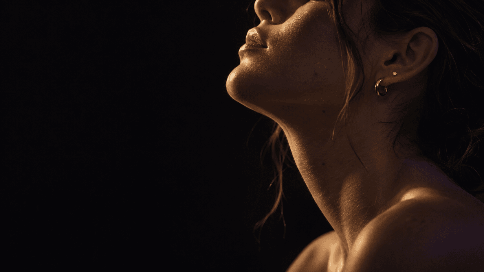 Perfil em close de uma pessoa com piercings de ouro, olhando para a luz, fundo escuro.