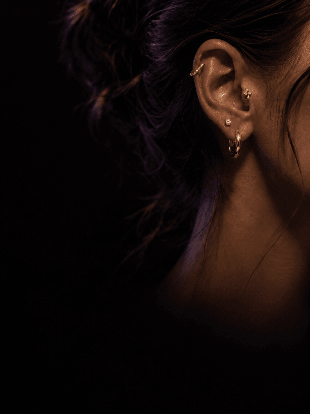 Close cinematográfico de uma orelha com piercings e joias em ouro, luz dramática lateral sobre fundo escuro.